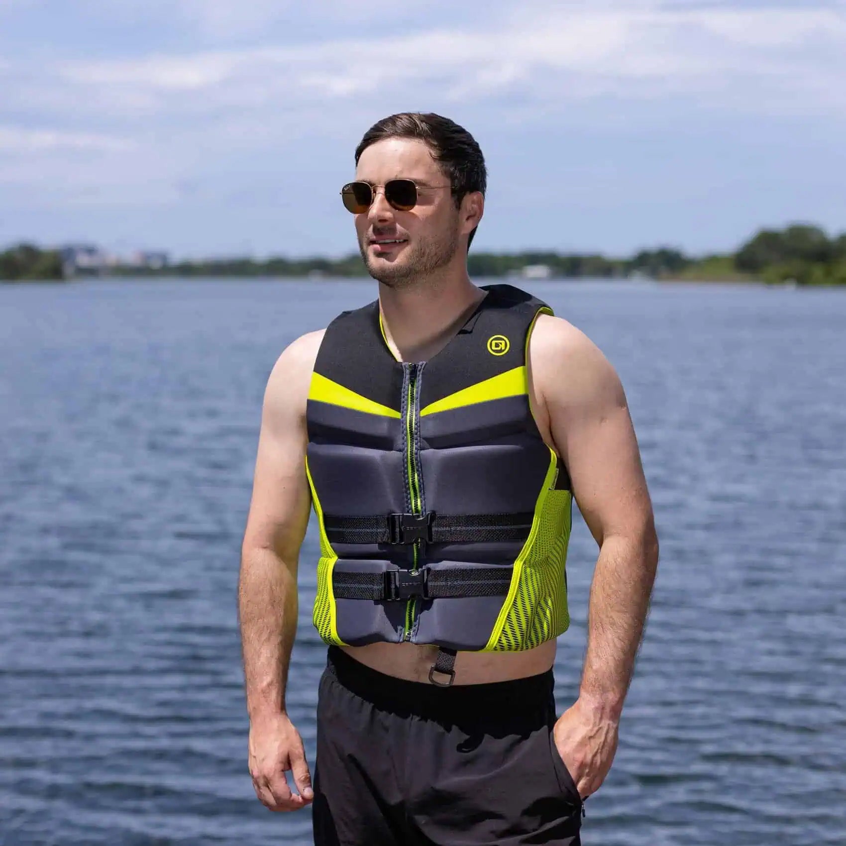 Man wearing a gray and yellow O'Brien Men's Flex V-Back Life Jacket standing by a body of water.