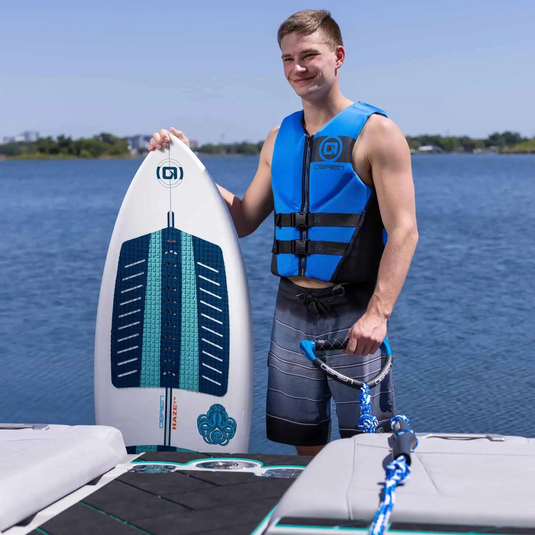 Person holding a wakeboard with a blue O'Brien Men's Traditional Vest on a dock by water