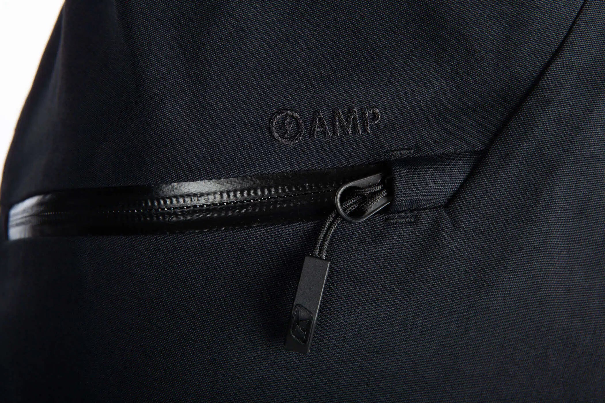 Close-up of a Klim Togwotee Bib showing a black zipper pocket with a black pull tab and an embossed AMP logo.