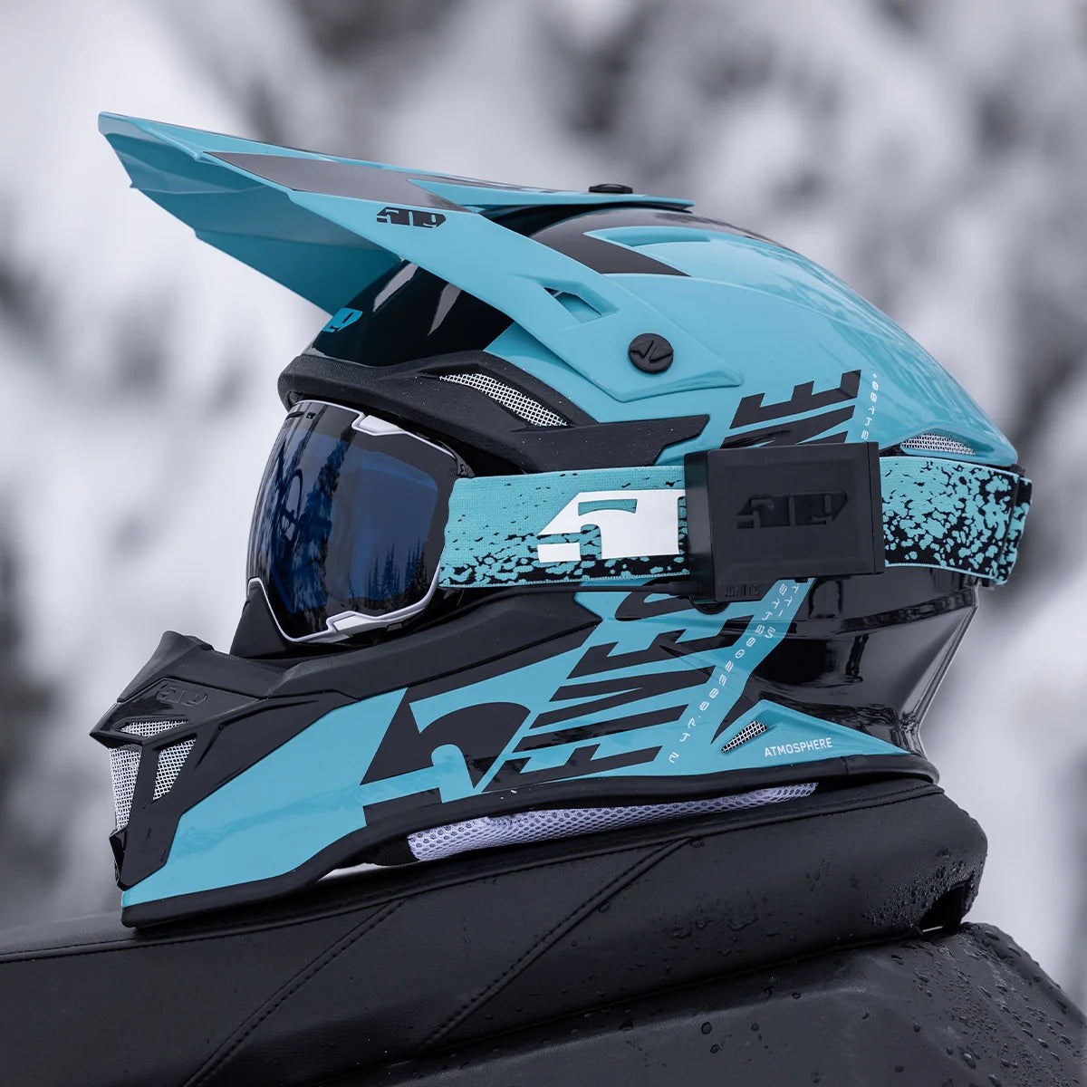 a 509 Atmosphere Snowmobile Helmet - Glacier Blue sitting on the back of a snowmobile with grey snowing blurred background