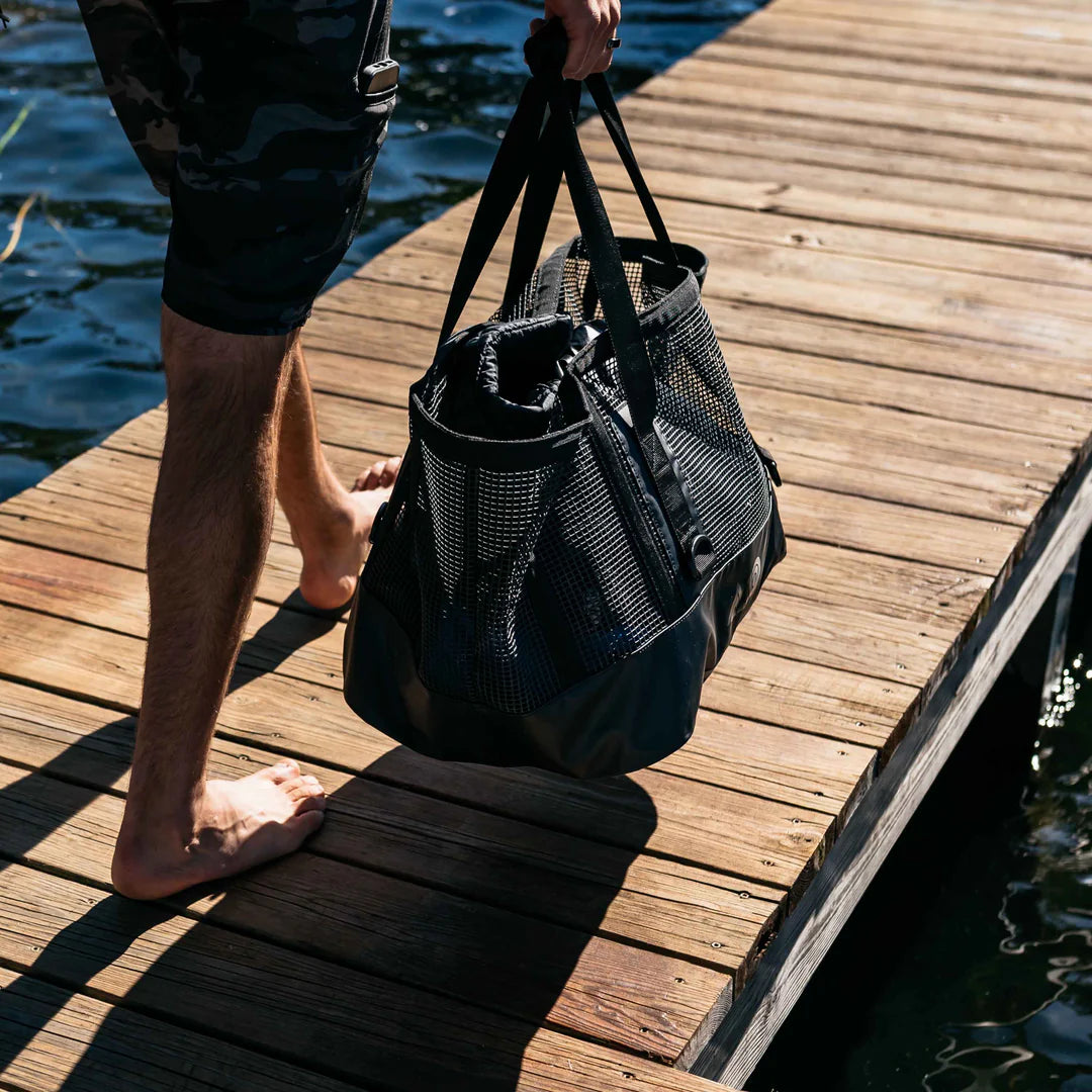 Person holding a black Mission Cassi Gear Tote - Heavy-Duty Boat and Beach Bag on a wooden dock by water