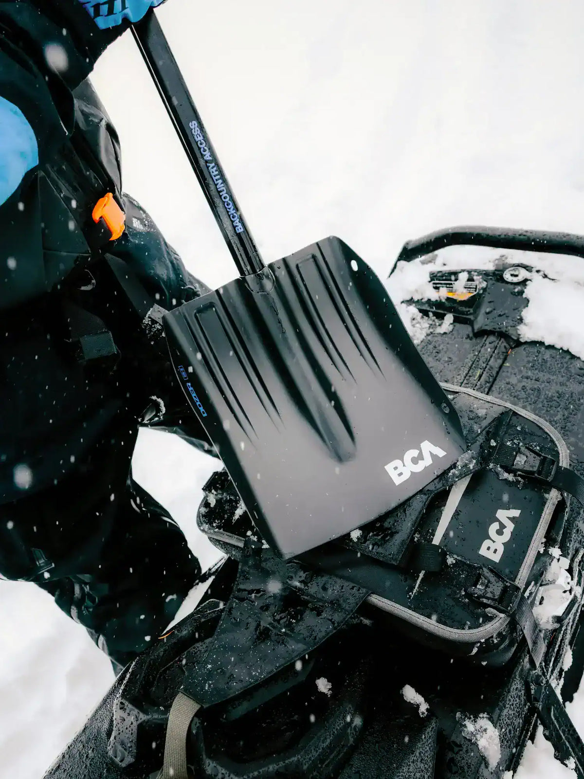 A close-up action shot of a BCA Dozer 2H-S Shovel with Folding Saw being used on a snowmobile in the snow, with a person's arm and body visible on the left.