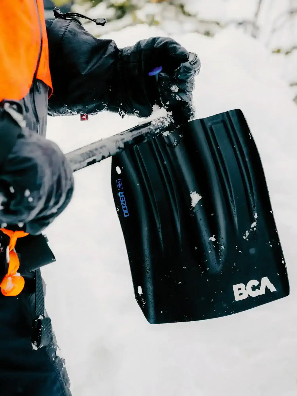 A close-up action shot of a person shoveling snow with a black BCA Dozer 2H-S Shovel with Folding Saw. The person is wearing winter gear, including an orange and black jacket and black gloves, in a snowy outdoor setting.