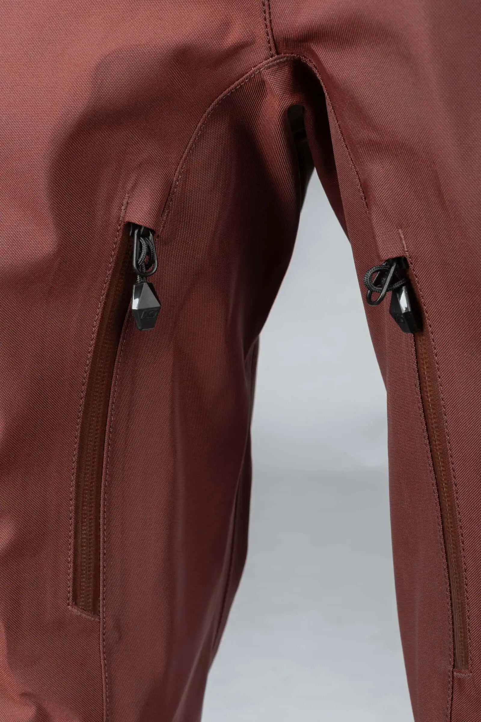 Close-up of Klim Lochsa One-Piece Monosuit - Bitter Chocolate - Montana Tan showing two zippered pockets with black zipper pulls against a light gray background.