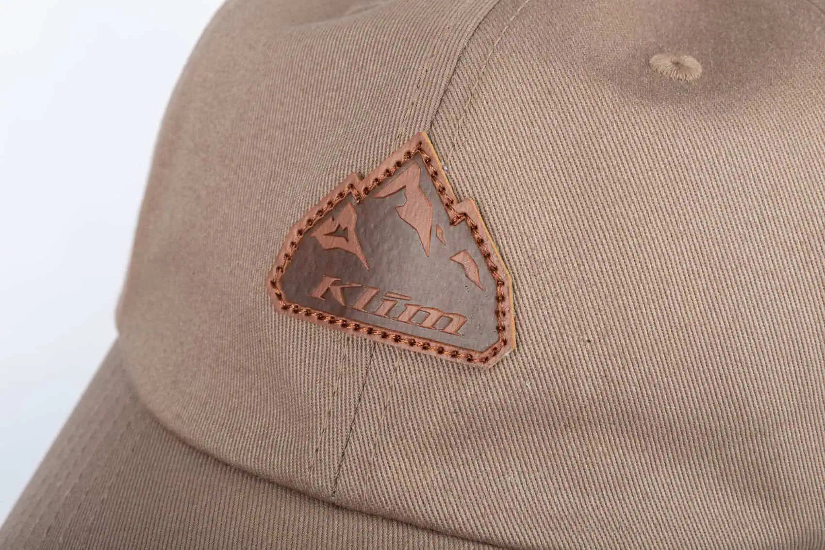 Close-up of a tan Klim Peak Hat featuring a brown stitched triangular patch on the front with the embossed brand name 'Klim'.