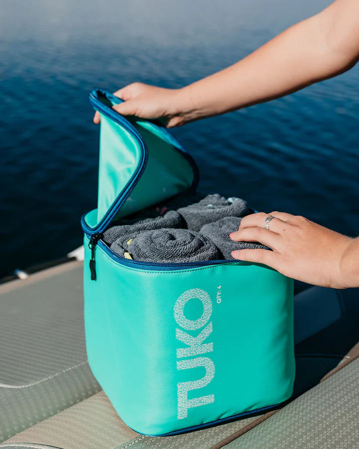Turquoise TUKO bag with gray towels being packed by a person by a body of water.