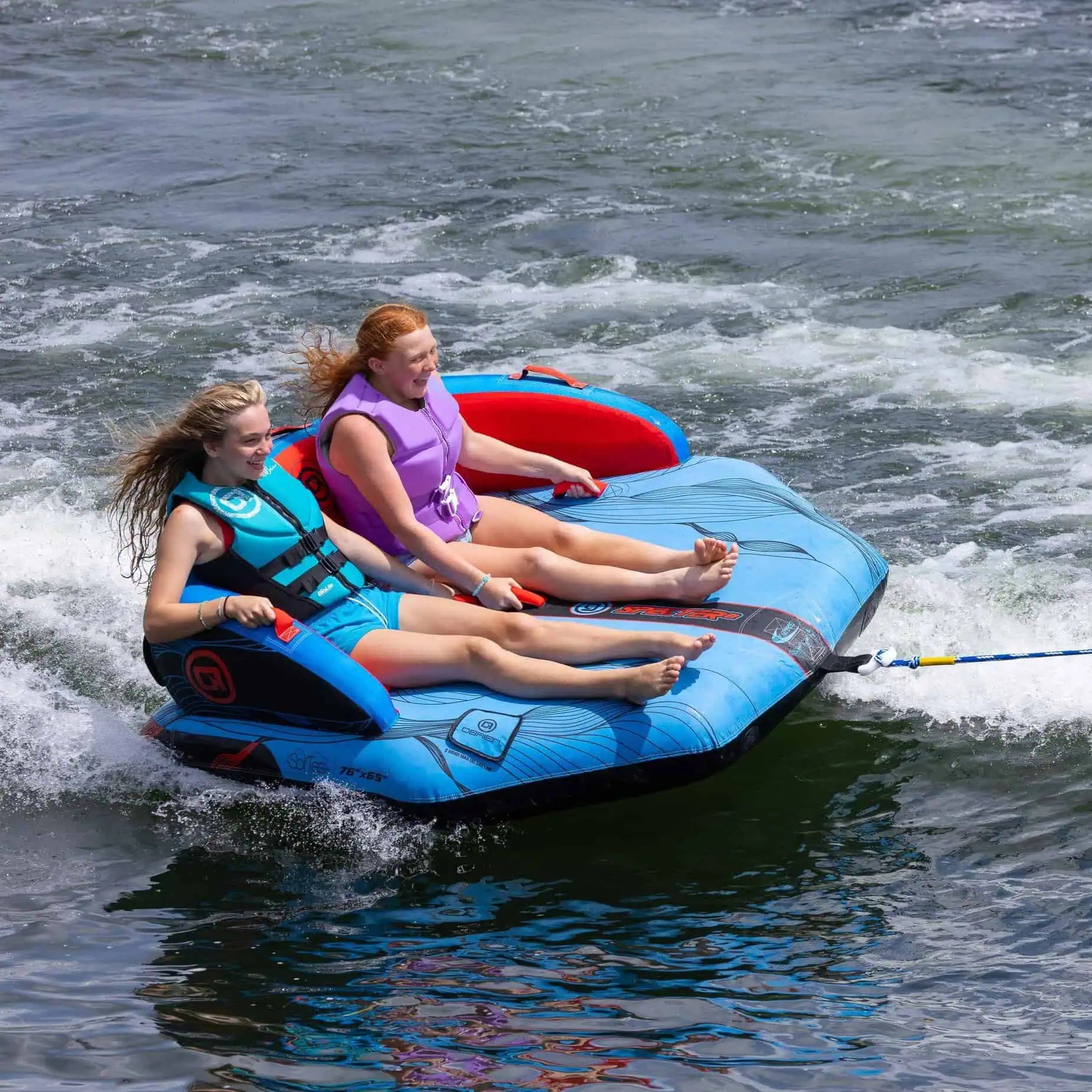 Two people on a blue and red inflatable O'Brien Specter 2 Boat Tube being towed through water.