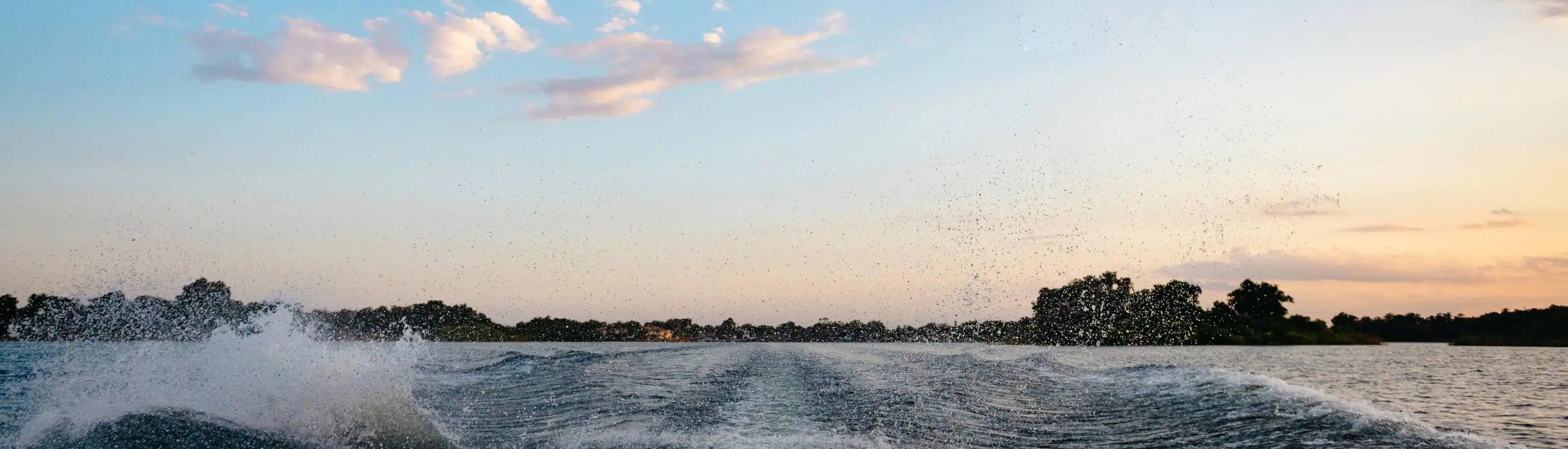 A scenic sunset over water with a wake from a boat, showing splashing white water, dark tree silhouettes along the horizon, and a blue sky with soft pink clouds.