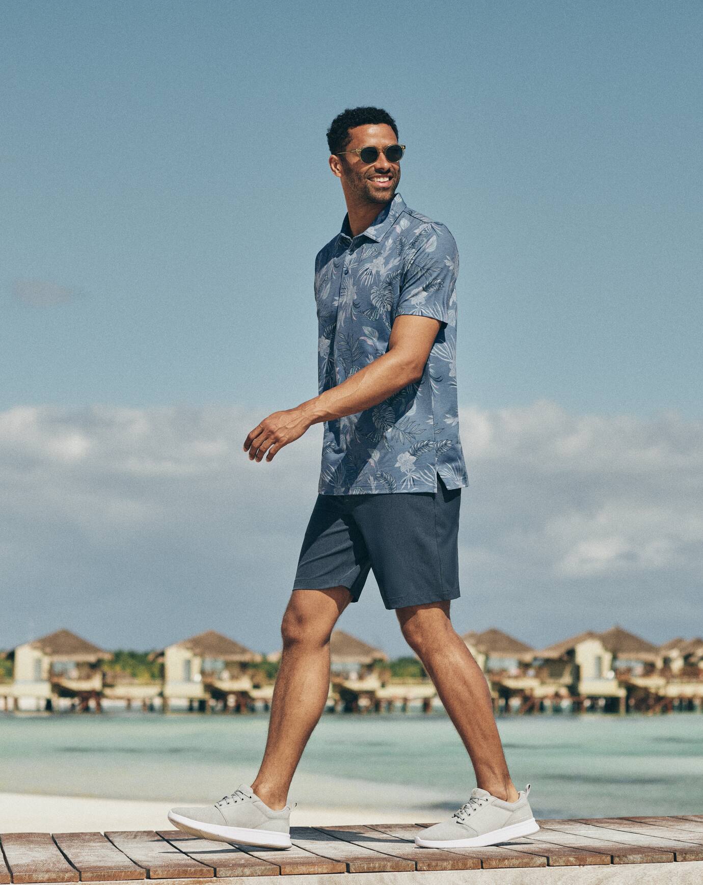 Man walking on a wooden deck by the water with a clear blue sky and distant buildings, wearing the TravisMathew Featherweight Reservation Polo Shirt