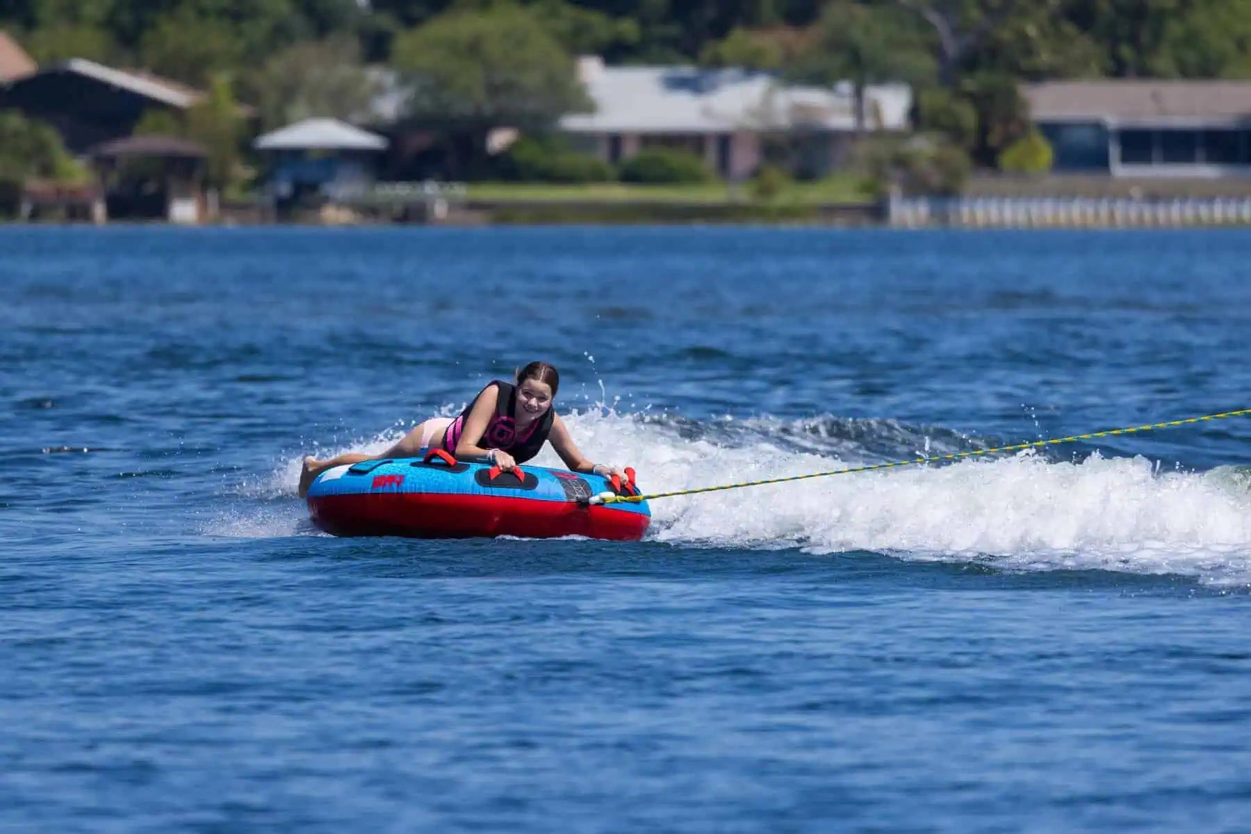 Person on a red O'Brien Screamer Tube being pulled by a rope on a lake with houses in the background