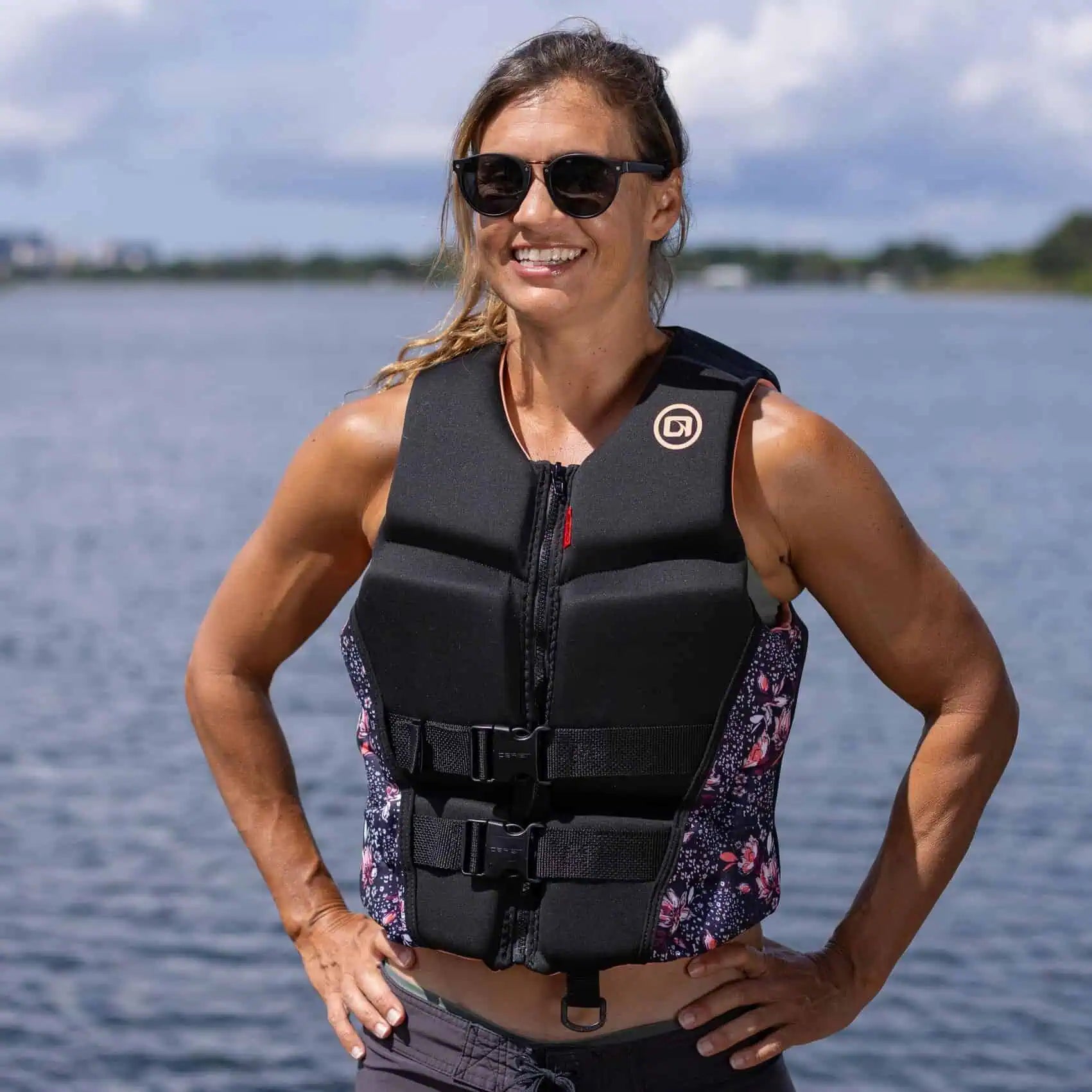 Woman wearing a coral/floral O'Brien Ladies Flex V-Back Life Jacket and sunglasses by a body of water