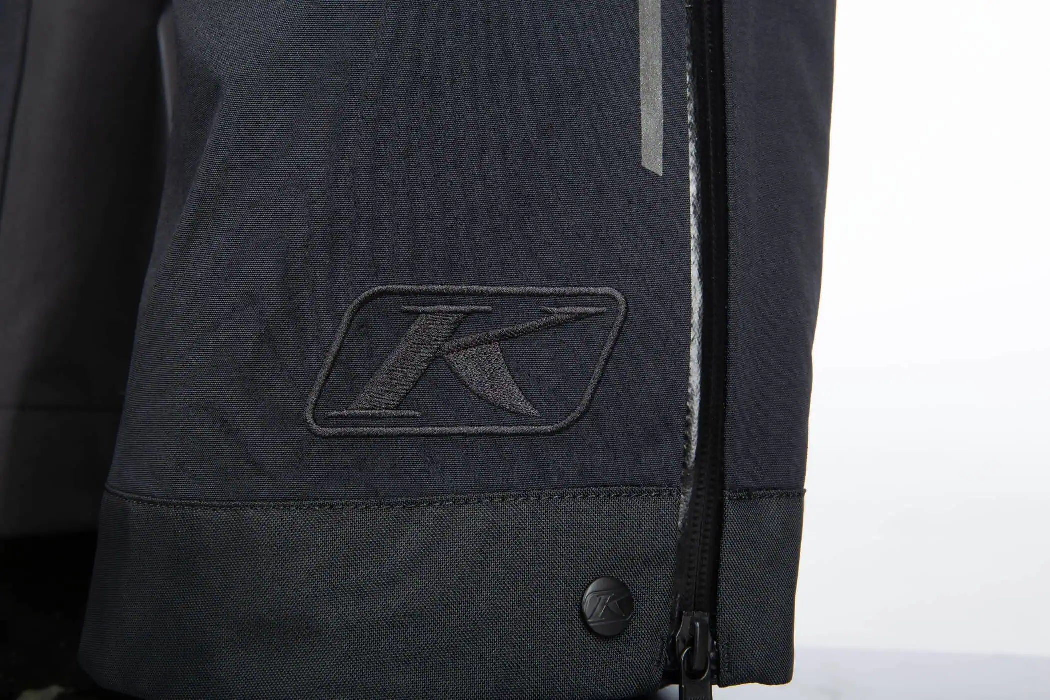 Close-up of a Klim Togwotee Bib cuff showing a black embroidered stylized 'K' logo patch and a vertical reflective strip near the zipper.