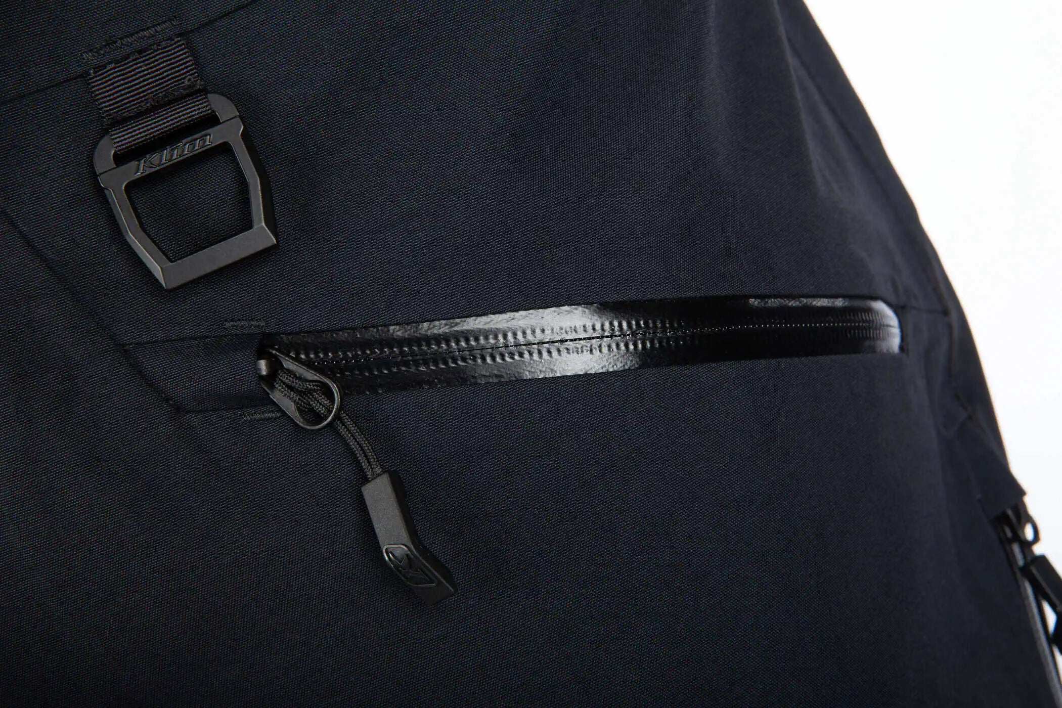 Close-up of a Klim Togwotee Bib showing a horizontal zippered pocket with a glossy black zipper pull and a black plastic buckle detail on the side.