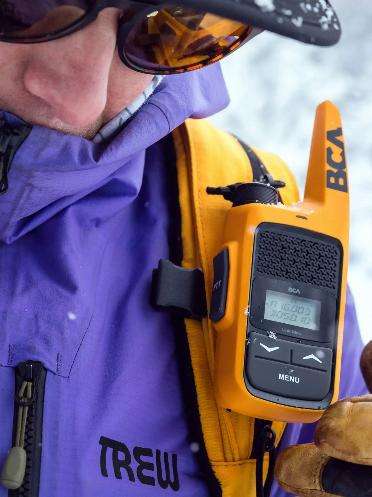 Person wearing a purple jacket with a yellow BCA BC Link Mini Radio on a snowy background
