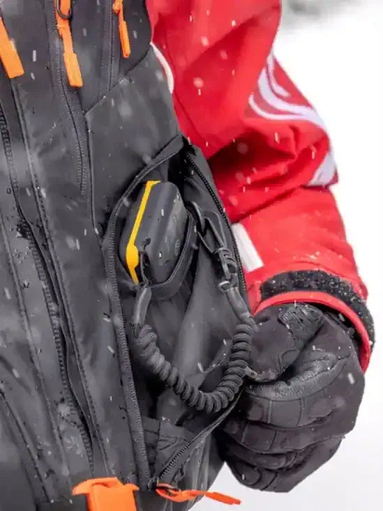 Person wearing a red jacket and black gloves holding a BCA Tracker3+ Avalanche Transceiver in a snowy environment.