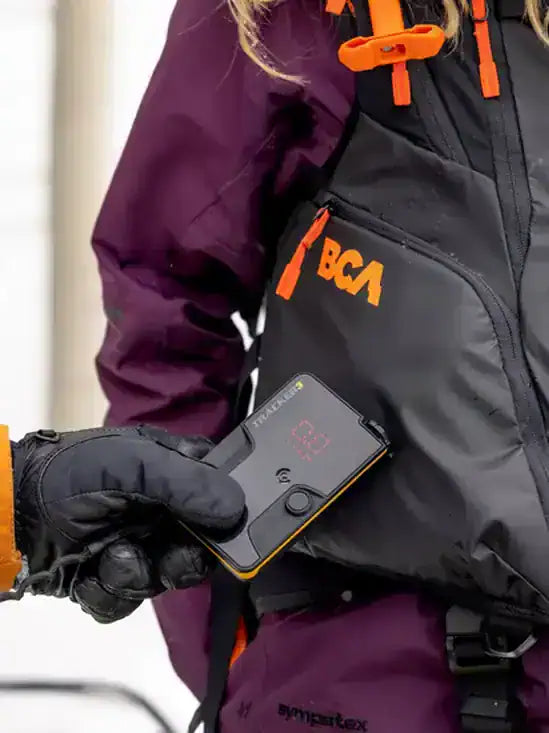 Person wearing a jacket with BCA branding holding a BCA Tracker3+ Avalanche Transceiver