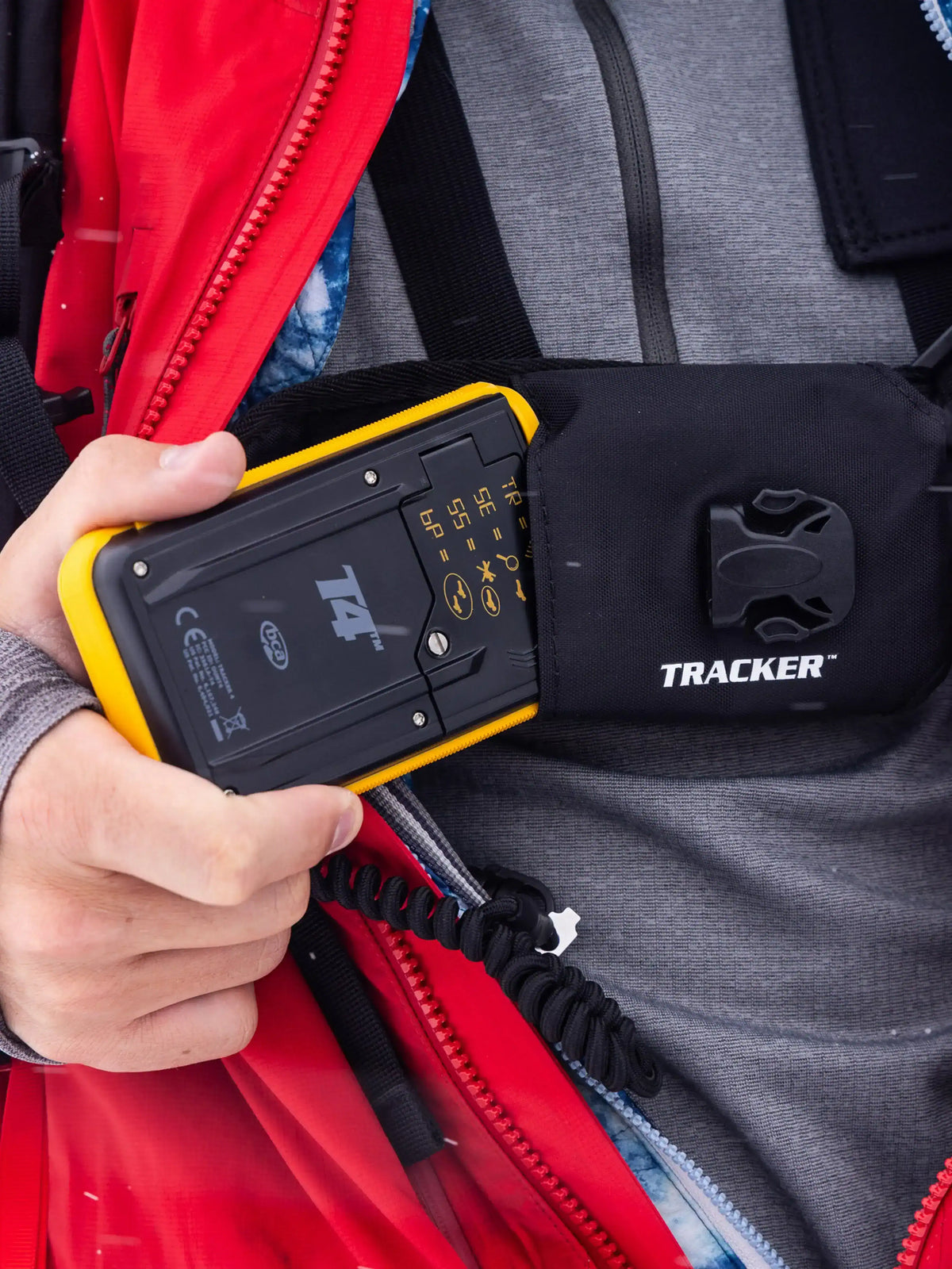 Hand holding a BCA Tracker4 Avalanche Transceiver against a red jacket and gray backpack.
