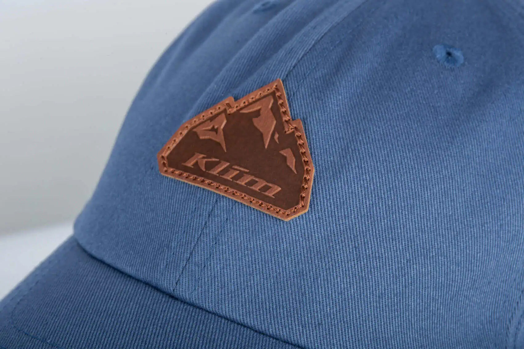 A close-up shot of a blue Klim Peak Hat featuring a brown leather patch on the front with the brand name 'Klim' and mountain graphic.
