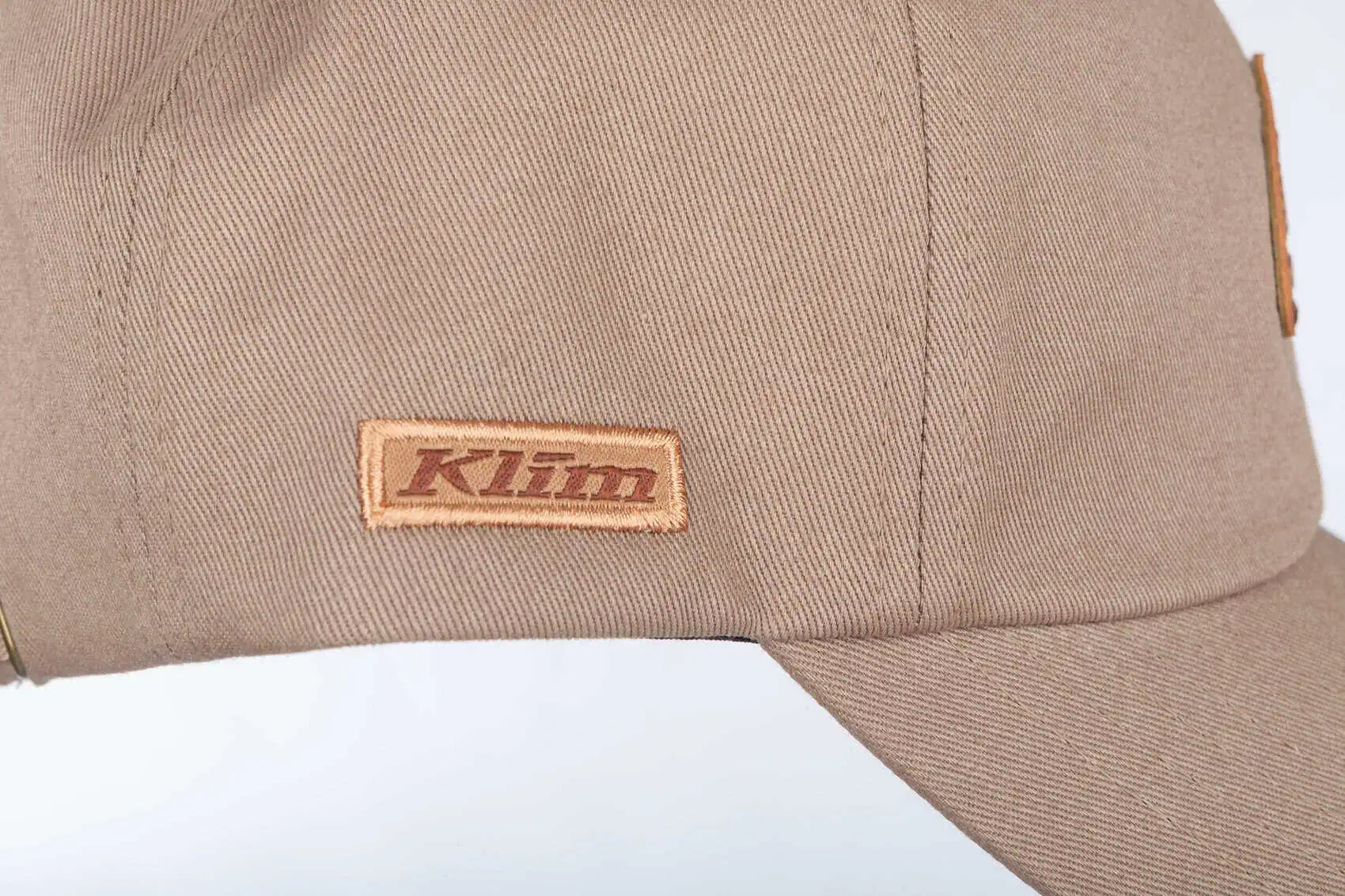 Close-up of a tanKlim Peak Hat showing the side panel with a rectangular brown Klim logo patch and part of the brim against a white background.