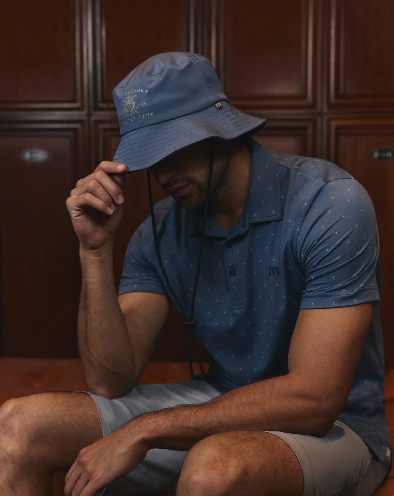 Man wearing a blue bucket hat and matching polo shirt sitting against wooden paneling.